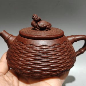 Può includere: Una teiera in ceramica marrone con un design a trama di vimini. Il coperchio presenta una piccola figura di animale scolpita. Il manico è a forma di bambù. La teiera è tenuta in una mano.