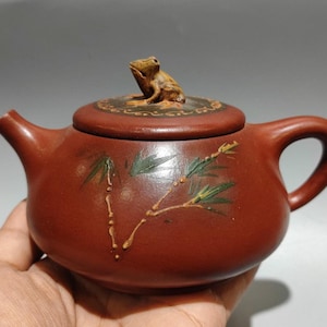 以下が含まれることがあります： 蓋にカエルの置物がある赤茶色の陶器のティーポット。 ティーポットは、曲がった注ぎ口とハンドルを備え、竹の葉と茎が描かれています。 ティーポットは手で持たれています。