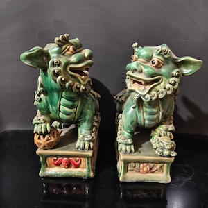 May include: A pair of green ceramic Foo Dog statues. Each statue has a fierce expression, detailed fur, and sits atop a rectangular base with floral and scrollwork designs. One holds a ball under its paw. The statues are approximately 10 inches tall.