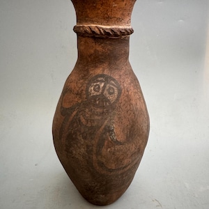 May include: A brown ceramic vase with a wide mouth and a narrow neck. The vase is decorated with a black and white design of a stylized human figure.