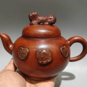 May include: A reddish-brown clay teapot featuring a rounded body, a curved spout, and a handle. The lid is topped with a sculpted animal figure. The exterior surface is decorated with three raised circular and heart-shaped medallions.
