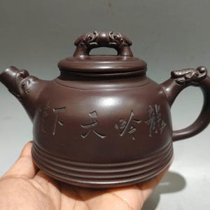 Könnte beinhalten: Braune Keramik-Teekanne mit Deckel und Henkel. Die Teekanne hat einen Ausguss und ist mit geschnitzten chinesischen Schriftzeichen verziert. Der Griff und der Deckel weisen drachenartige Designs auf.