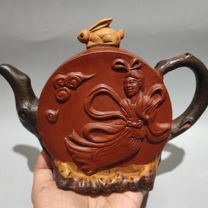 May include: A reddish-brown clay teapot featuring a relief carving of a figure in flowing robes amidst clouds. The teapot has a dark brown handle and spout resembling tree branches, and a small sculpted rabbit figure sits atop the lid.