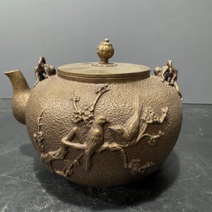 Könnte beinhalten: Eine antike Teekanne aus Messing mit einer strukturierten Oberfläche und aufwendigen Vogel- und Blumenmotiven. Die Teekanne hat einen Deckel mit einem Knauf und zwei Griffen.