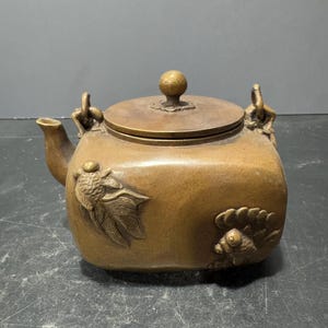 May include: A square-shaped brass teapot with a lid and two handles. The teapot is decorated with raised fish designs.