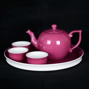 May include: A magenta tea set with a teapot, three cups, and a serving tray. The teapot and cups are magenta with white interiors. The tray is magenta with a white rim. The set is on a black background.