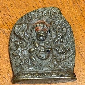 May include: A bronze-colored, religious figurine depicting a seated deity with multiple arms and a crown. The figure is surrounded by a flame-like design. The figurine is on a wooden surface.