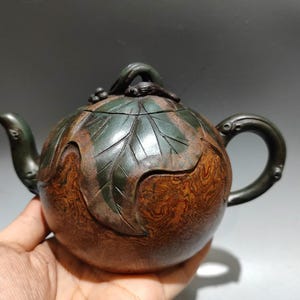 以下が含まれることがあります： 手作りのアンティークティーポット。濃い茶色と緑色の仕上げ。葉のような彫刻が施された丸いボディと湾曲したハンドルが特徴です。注ぎ口は湾曲しており、蓋には小さなノブが付いています。ティーポットは手で持たれています。