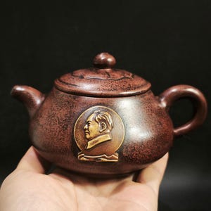 Könnte beinhalten: Eine braune Keramik-Teekanne mit Deckel und Griff. Die Teekanne zeigt ein kreisförmiges Relief eines Männerprofils auf der Vorderseite. Die Teekanne wird in einer Hand vor dunklem Hintergrund gehalten. Die Teekanne ist im traditionellen Stil gehalten.