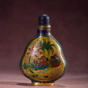 May include: An ornate, antique cloisonné snuff bottle. The bottle features a yellow background with a detailed design of two birds, a lotus flower, and foliage. The bottle has a dark blue stopper and gold accents.