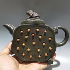 Puede incluir: Tetera de cerámica verde oscuro con una figura de rana en la tapa. La tetera tiene un diseño único con múltiples hendiduras circulares llenas de acentos marrón claro. El asa y el pico son curvos.