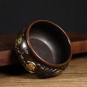 Könnte beinhalten: Ein kleiner, runder, dunkelbrauner Räucherofen mit aufwendigen Schnitzereien und goldenen Akzenten. Der Ofen hat eine kreisförmige Öffnung oben und ist mit chinesischen Schriftzeichen verziert.