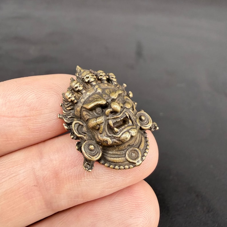 May include: A detailed bronze-colored metal pendant in the shape of a fierce face with a wide open mouth and multiple skulls on top. The pendant has intricate details and a weathered appearance.