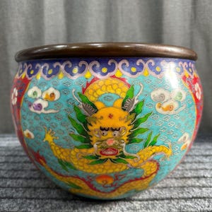 Peut inclure: Un bol rond décoratif en cloisonné avec un bord en bronze. Le bol présente un fond turquoise avec un motif de dragon jaune. Des motifs floraux rouges et blancs et des nuages blancs sont également présents. Un objet d'art décoratif.