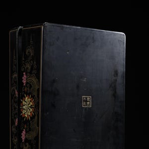 May include: A rectangular black lacquer box featuring gold and red floral motifs along the side panel. The front surface displays a small square seal with the Chinese characters 乾隆年製 in gold. The box has a smooth, dark finish and sharp edges.