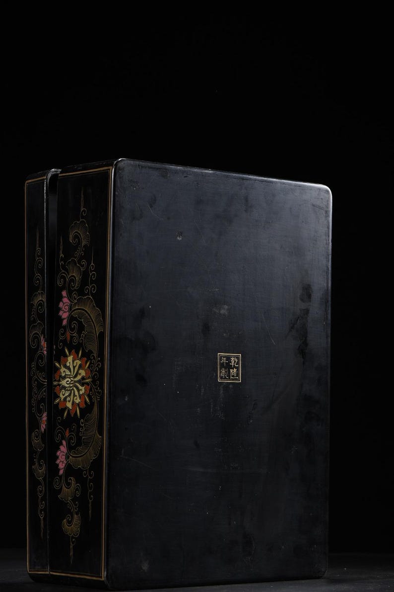 May include: A rectangular black lacquer box featuring gold and red floral motifs along the side panel. The front surface displays a small square seal with the Chinese characters 乾隆年製 in gold. The box has a smooth, dark finish and sharp edges.