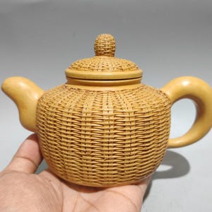 May include: A light tan teapot with a textured surface that mimics the appearance of woven wicker. The teapot features a rounded body, a curved spout, a smooth handle, and a matching lid with a small, textured knob on the top center.