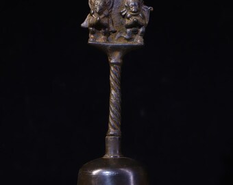 May include: A bronze bell with a twisted handle and a decorative top featuring two figures. The bell has a flared base and a dark, aged appearance. The top has a stylized design, possibly religious or symbolic.