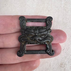 May include: A dark bronze-colored buckle with an ornate design. The buckle features a stylized face with detailed features and scrollwork. The rectangular buckle has two openings for attachment and is held in a hand.