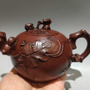 Puede incluir: Tetera de cerámica marrón con intrincadas tallas de flores y hojas. La tetera presenta un asa y un pico curvos, y una tapa con un pequeño elemento decorativo. El diseño es detallado y elegante.