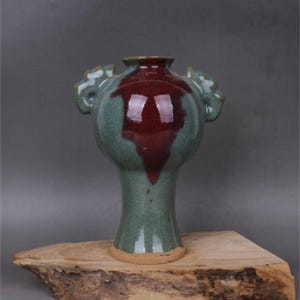 May include: A green and red glazed ceramic vase with two handles. The vase has a round body and a tall, slender neck. The vase is sitting on a wooden surface.