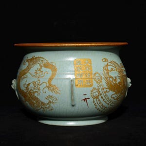 May include: A light blue ceramic bowl with gold dragon and phoenix designs. The bowl has a gold rim and handles. Chinese characters are visible on the bowl. The bowl is on a black background.