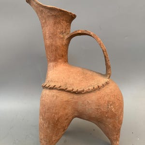 May include: A terracotta pottery jug with a tall, flared spout and a handle. The jug has three legs and a decorative band around the body.