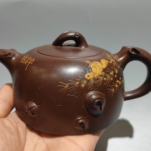 以下が含まれることがあります： 曲線的なハンドルと注ぎ口が付いた茶色のセラミックティーポット。 ティーポットは小さなループが付いた蓋を備え、ネズミと葉の金色のイラストで装飾されています。 ティーポットの側面にはテキストがあります。
