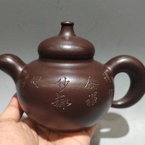 以下が含まれることがあります： 丸みを帯びたボディ、湾曲した注ぎ口、ループハンドルを備えた茶色のセラミックティーポット。蓋には小さなノブが付いています。ティーポットの本体には漢字が刻まれています。ティーポットは濃い茶色です。