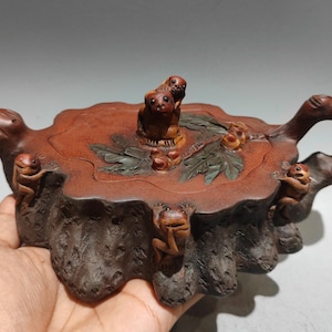 Puede incluir: Una tetera de cerámica marrón con forma de tocón de árbol y exterior de corteza texturizada. La tapa presenta figuras esculpidas de pequeños animales y hojas verdes. Otras figuras de animales están adheridas a los lados del cuerpo.
