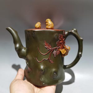 Pode incluir: Uma chaleira artesanal com um design de tronco de árvore. A chaleira verde escura apresenta uma alça e um bico de galho, com macacos e folhas esculpidas. A tampa tem duas pequenas figuras de macaco. A chaleira é um item decorativo.