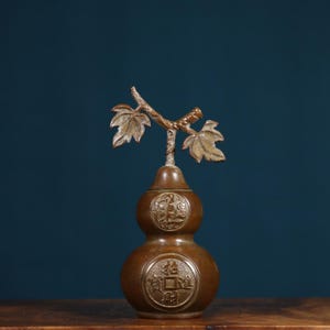 May include: A brown gourd-shaped vase with a decorative vine and leaves on top. The vase has two round sections with Chinese characters embossed on them.