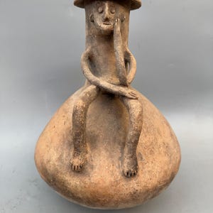 May include: A terracotta sculpture of a seated figure with arms crossed and a hand touching the face. The figure has a cylindrical head with a hat and is seated on a rounded base. The sculpture is a light brown color.