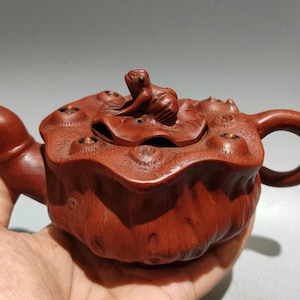 May include: A handcrafted, reddish-brown teapot carved from wood. The teapot features a frog-shaped lid and a lotus flower design. The spout and handle are curved. The teapot is held in a hand.
