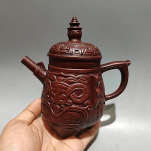 May include: A dark brown, intricately carved teapot with a lid and handle. The teapot features detailed relief carvings across its body and lid. The spout is angled, and the handle is curved. The teapot is held in a hand.