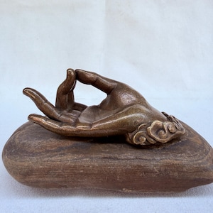 May include: Bronze-colored sculpture of a hand in a mudra pose, resting on a brown, oblong base. The hand's details are finely sculpted, with a focus on the fingers and the gesture. The base has a smooth, rounded shape.