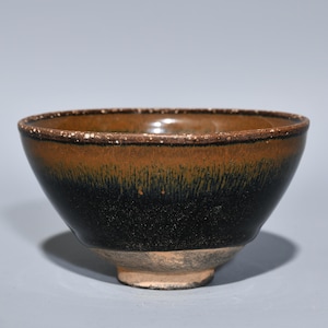 May include: A ceramic bowl with a dark brown and black gradient glaze. The bowl has a rounded shape with a slightly flared rim and a small foot. The glaze has a textured appearance, and the bowl appears to be antique.