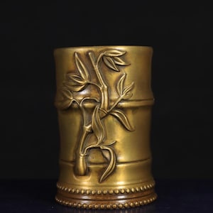 May include: A cylindrical, gold-colored metal container with a bamboo design. The bamboo stalk and leaves are raised from the surface. The base has a beaded design. The container is likely used for holding items.