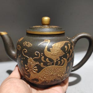 May include: Dark brown teapot with gold accents. The teapot features a crane and dragon design. The lid and spout are also accented with gold. The handle is curved and the teapot is held in a hand.