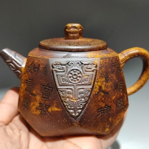 May include: A brown, antique-style teapot with a lid and handle. The teapot features intricate carvings and Chinese characters. The body of the teapot has a shield-shaped design in the center. The spout is angled and the handle is curved.