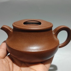 May include: A small, brown ceramic teapot with a rounded body, a curved spout, and a circular handle. The lid has a small opening. The teapot is held in a hand against a neutral background. The teapot is likely used for brewing tea.