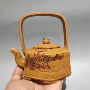Puede incluir: Tetera cuadrada de color marrón claro con asa curva y boquilla. La tetera presenta un diseño de paisaje con árboles, montañas y una pagoda en tonos marrones y rojos. La tapa tiene un pequeño pomo redondo.