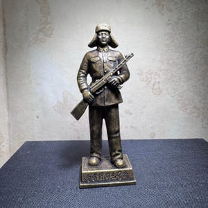May include: A bronze statue of a soldier wearing a fur hat and holding a rifle. The statue is standing on a base with Chinese characters.