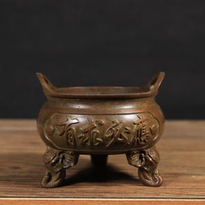 May include: A bronze-toned metal incense burner featuring a rounded body with two upright handles and three legs shaped like elephant heads. The exterior surface displays embossed Chinese characters that read 有求必應, indicating a prayer for fulfillment.