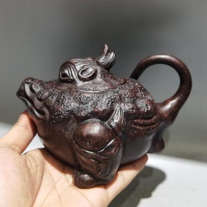 May include: A dark brown, intricately carved teapot in the shape of a frog. The teapot features a detailed textured surface, a curved handle, and a small spout. The frog's face is prominent, with detailed eyes and a smiling mouth. The lid is also carved.