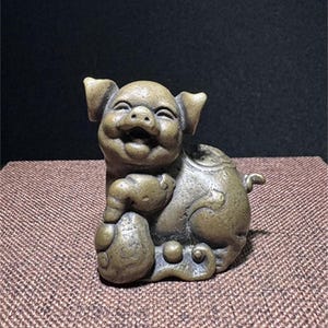 May include: A small, bronze figurine of a smiling pig holding a round object in its paws. The pig is sitting on a textured surface.