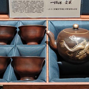May include: A dark brown clay tea set consisting of a teapot with a gold-coloured dragon relief and four matching cups, presented in a wooden box lined with dark blue fabric. An informational card with Chinese text is included in the lid.