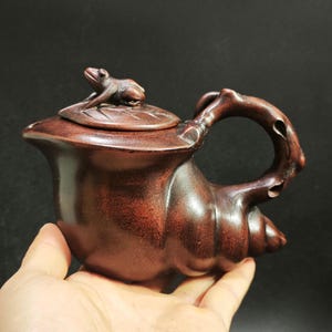 Puede incluir: Tetera de madera marrón rojiza hecha a mano con una figura de rana en la tapa. La tetera tiene una forma única y orgánica con un asa curvada y un pico que se asemeja a una concha. La tapa tiene forma de hoja.