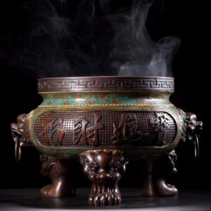 Op de afbeelding: Een bronzen wierookbrander met een turquoise en rood bloemmotief, ingewikkelde gravures en drakenkopgrepen. Wispelturige rook stijgt op uit de top tegen een zwarte achtergrond. De brander rust op drie sierlijke leeuwenpootvoeten.