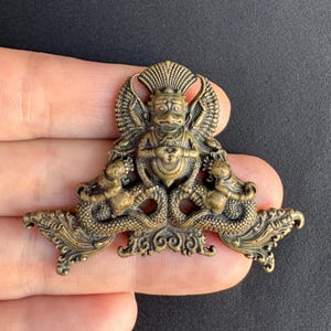 May include: A bronze-colored religious pendant featuring a central figure with a headdress, flanked by two serpent-like creatures. The pendant has intricate details and a decorative design, likely for spiritual or ornamental purposes.